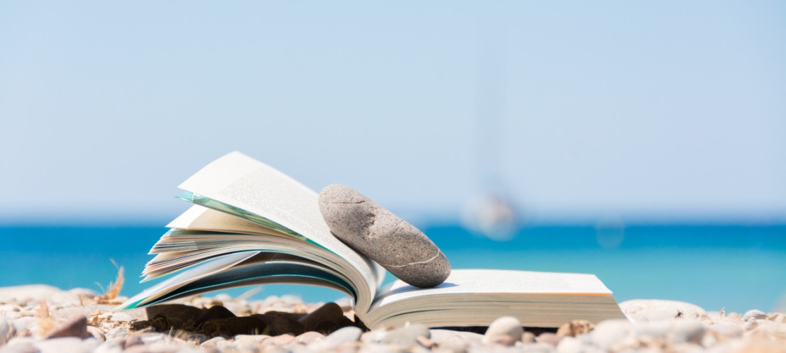 Een boek ligt op het strand waarbij een steen het boek openhoudt bij de pagina waar de vakantielezer is gebleven.